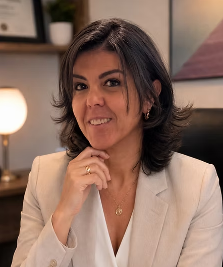 Karina Ovalle em seu consultório, de blazer branco, apoiando o queixo na mão.
